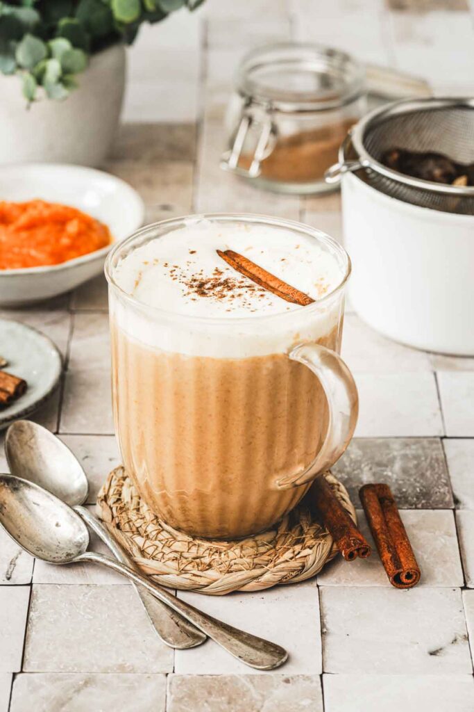 Tasse en verre remplie de chai latte doré, garnie de crème blanche à la vanille et de poudre et d'un bâton de cannelle. À côté, on retrouve deux cuillères en métal et un bâton de cannelle et, en arrière-plan, un bol blanc avec un ingrédient rouge dedans, le petit pot Mason de cannelle et un autre contenant blanc.