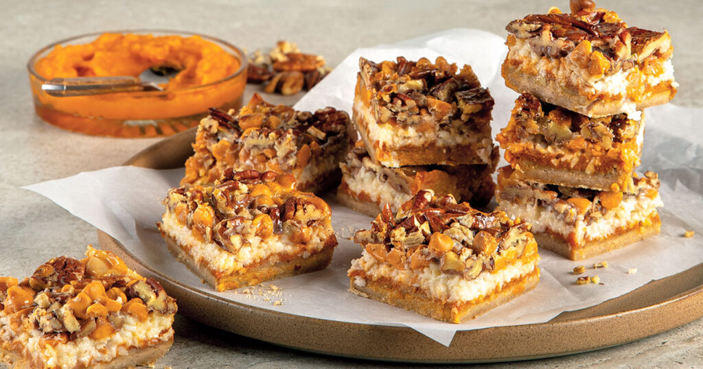 Carrés à la citrouille empilés sans un plateau sur une feuille de papier ciré. Huit morceaux sont dans le plateau et un est seul en bas à gauche de l'image. En arrière-plan, il y a une purée à la citrouille dans un bol avec une cuillère.