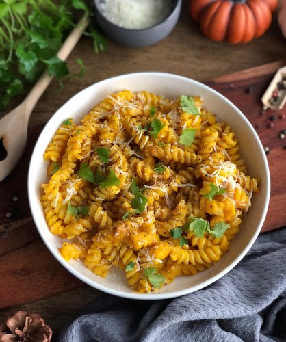 Bol de pâtes à la citrouille avec pecorino et parmesan avec les deux ingrédients en arrière-plan et une citrouille. Il y a également un linge à vaisselle en bas de l'image.