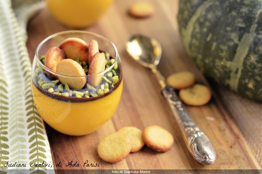 Panna cotta à la citrouille dans un petit pot en verre, garnie d'une mince couche de chocolat, de pistache et de cinq petits biscuits à la vanille ronds. Sur la table où repose le pot, il y a six autres petits biscuits répandus, une cuillère en métal et une serviette blanche avec des motifs de feuilles dessus.