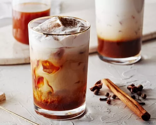 Café infusé à froid avec de la crème à la citrouille et saupoudré de cannelle. Deux autres cafés sont en arrière-plan, avec un bâton de cannelle.