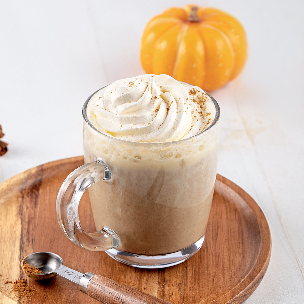 Latté à la citrouille dans une tasse transparente, avec de la crème fouettée sur le dessus et saupoudré de cannelle. La tasse est sur un plateau rond en bois avec une demi-cuillère à thé à mesurer, avec une citrouille en arrière-plan.