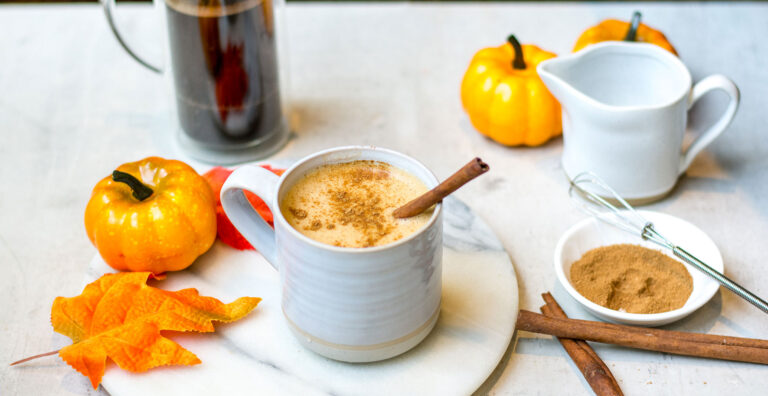 Chaï latté à la citrouille dans une tasse blanche, saupoudré de cannelle avec un bâton de cannelle au-dessus. La tasse est sur un plateau circulaire marbré blanc, avec une petite citrouille et deux feuilles d'automne. Autour du plateau se retrouvent de la cannelle avec un fouet, des bâtons de cannelle, deux autres petites citrouilles oranges, une tasse blanche et une tasse avec le mélange à café.