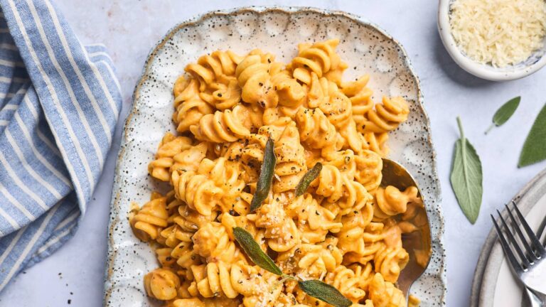 Demi-assiette rectangulaire de pâtes à la citrouille avec du poivre et des feuilles de sauge. Autour, on peut voir un petit bol de parmesan râpé, une serviette bleu et blanche lignée et le bout d'une fourchette.