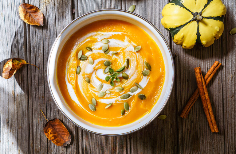 Bol de potage à la citrouille avec un tourbillon de crème et saupoudré de graines de citrouilles. Autour du bol se retrouvent des feuilles brunies par l'automne, deux bâtons de cannelle puis une citrouille verte et jaune.
